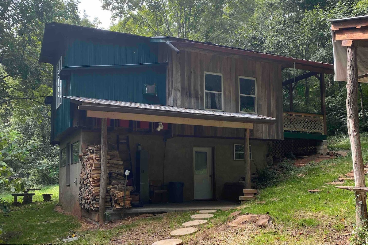 A rustic cabin is set amidst a woodland area, featuring a combination of wood and metal siding. Stacked firewood is positioned near the entrance, while a covered porch extends from the front, providing a space to enjoy the natural surroundings.