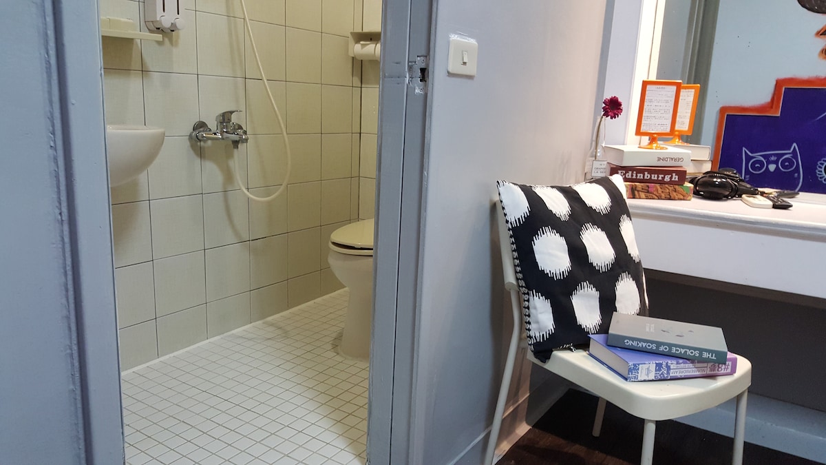 An interior view captures a compact bathroom area featuring a shower and toilet. A white chair with a black polka dot cushion is positioned near a small desk adorned with books and a decorative flower. Soft lighting enhances the simple decor.