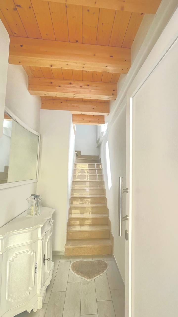 A light-filled hallway features a staircase leading to the upper level. The wooden ceiling beams add warmth to the space. A decorative console table is placed beside a mirror, and a heart-shaped rug is positioned at the foot of the stairs.