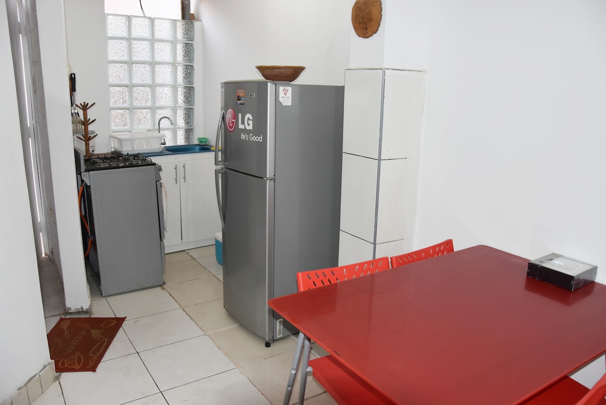 A compact kitchen area is visible, featuring a silver refrigerator and a four-burner gas stove. A small dining table with red chairs accommodates four people. The space is bright, with natural light coming through a window with glass blocks, and tiled flooring adds to the aesthetic.