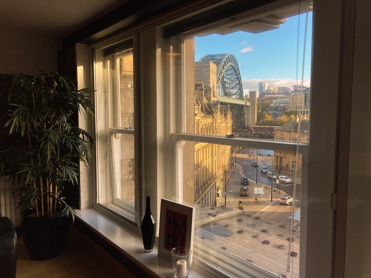 Large windows frame an iconic view of the Tyne Bridge and the bustling city below. A potted plant adds a touch of greenery to the interior, while artistic elements are displayed on the windowsill. Natural light fills the space, enhancing the inviting atmosphere.