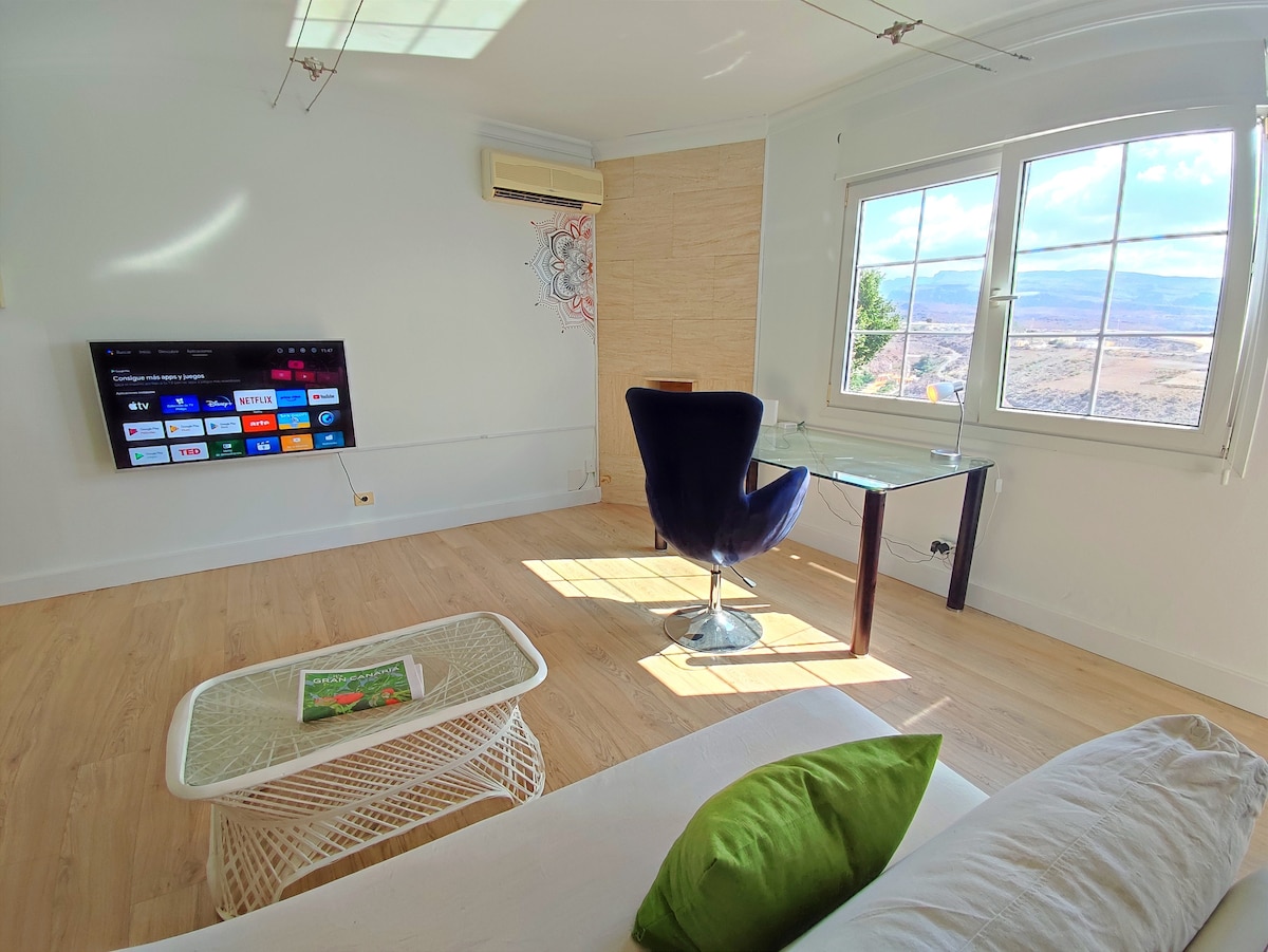 A bright living space features a comfortable chair and a glass desk, with a Smart TV mounted on a white wall. Natural light floods in through a large window, revealing panoramic views of the mountains, while a light-toned wooden floor adds warmth to the area.