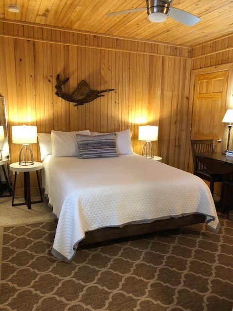 A cozy sleeping area features a queen bed with a textured white coverlet, complemented by two nightstands with lamps. The walls are lined with warm wood paneling, and a decorative fish is mounted above the bed. A small desk and chair are positioned nearby.