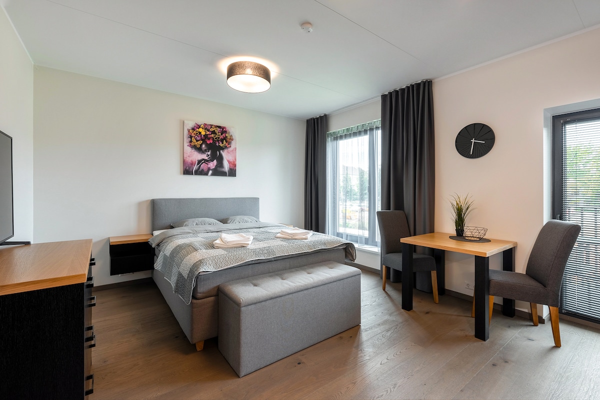 A modern bedroom features a spacious bed with a light grey headboard and a decorative plaid blanket. A small table and two chairs are positioned beside a large window, framed with dark curtains. A clock and a plant add subtle accents to the room.