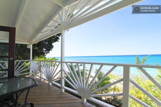 A spacious balcony features a wooden railing with a unique geometric design, offering unobstructed views of the Caribbean Sea. The scene is enhanced by lush greenery, creating a serene atmosphere. Sunlight reflects off the water, highlighting the refreshing blue tones of the ocean.