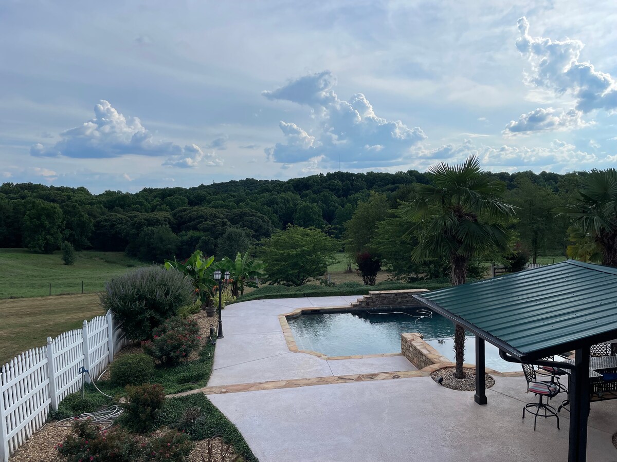 A serene backyard is visible, featuring a saltwater pool surrounded by a concrete deck. Lush greenery frames the area, with trees and open fields in the background. A shaded seating area is included, providing space for relaxation amidst the scenic landscape.