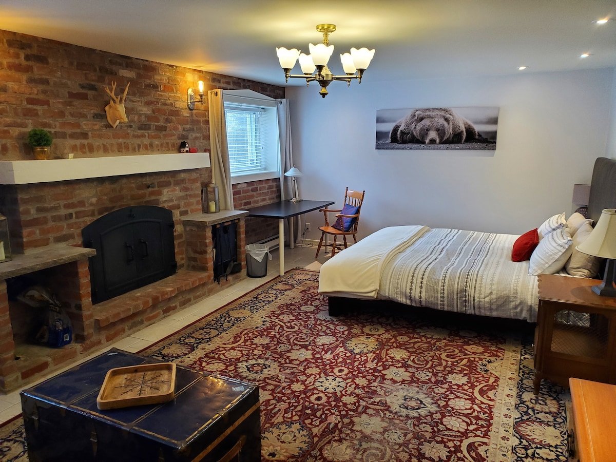 A spacious bedroom featuring exposed brick walls and a cozy fireplace. The room includes a large bed with soft linens, a vintage wooden rocking chair, and a desk. Light fixtures provide warm illumination, while a patterned rug adds comfort underfoot.