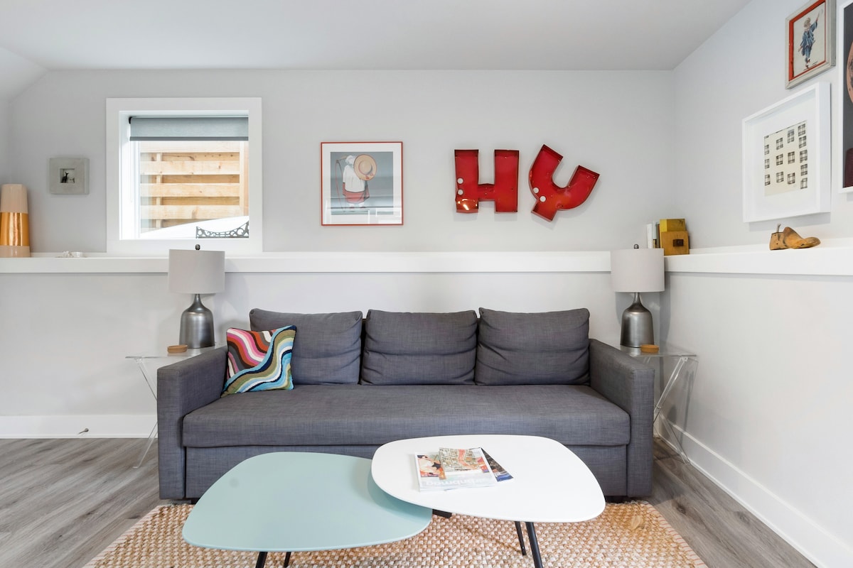 A modern living area features a sleek gray sofa with decorative cushions, positioned beside two stylish side tables. Framed artwork and vibrant wall decor are displayed above the sofa, complemented by warm lighting from unique lamps on either side.