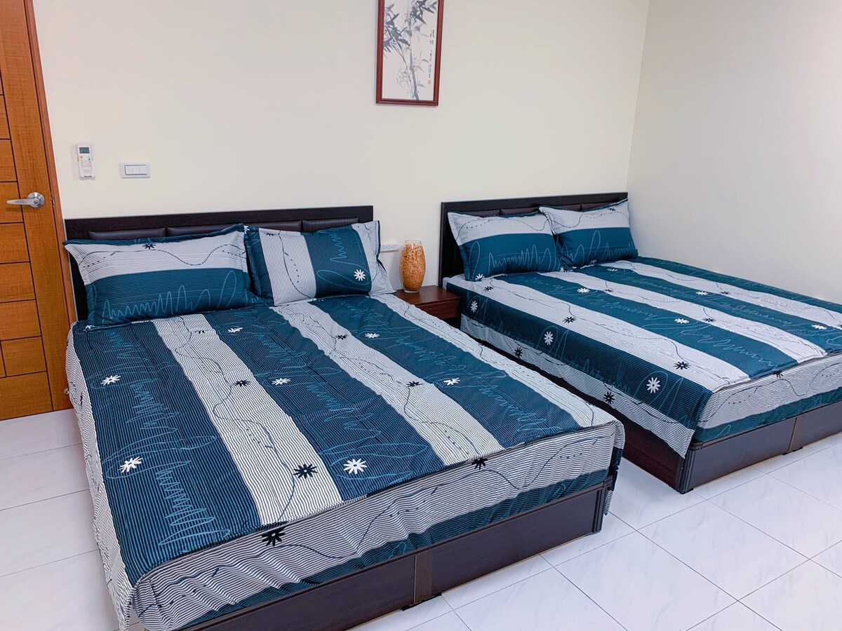 The sleeping area features two spacious beds, each outfitted with coordinated patterned bedding in shades of blue and white. A wood-framed artwork hangs above, complementing the neutral wall color. Natural light is present, enhancing the room's bright atmosphere.