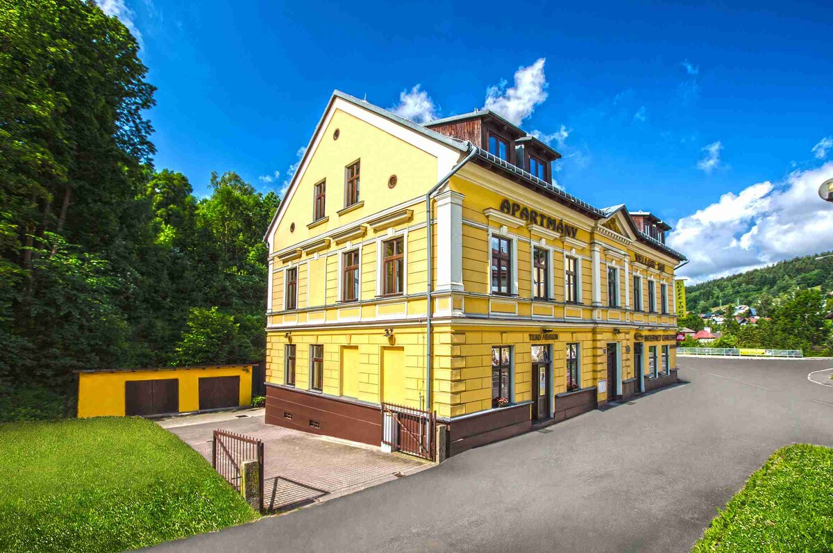 A charming three-story yellow building is shown, featuring classic architectural details. Surrounding greenery enhances the pleasant appearance. A spacious area is visible in the foreground, with a driveway leading to a garage. The clear blue sky adds to the inviting ambiance.