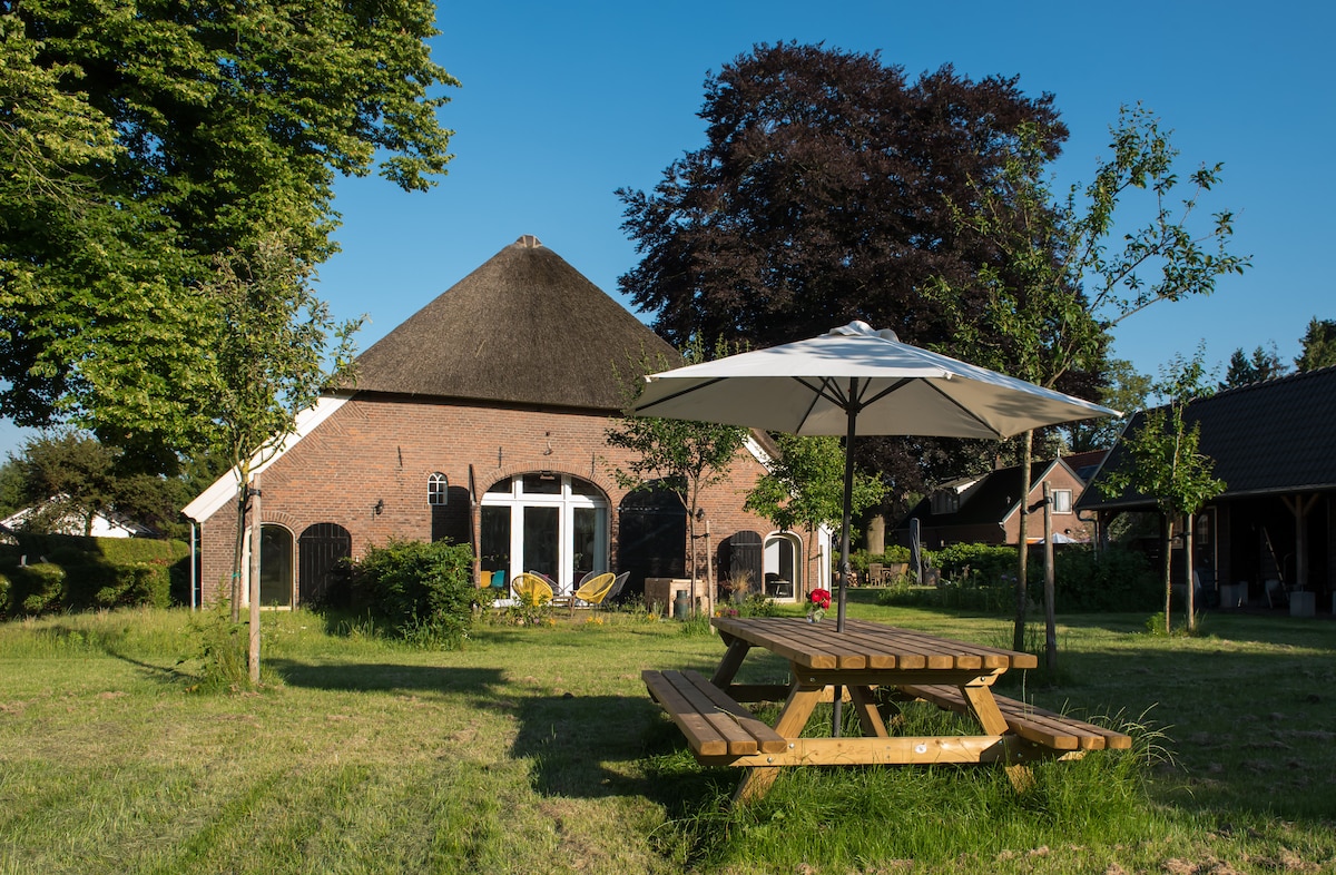 A thatched-roof cottage is surrounded by lush greenery. A large picnic table sits under an umbrella on the grassy area. Chairs and tables are visible beside expansive glass doors, leading into the inviting living space of the house.