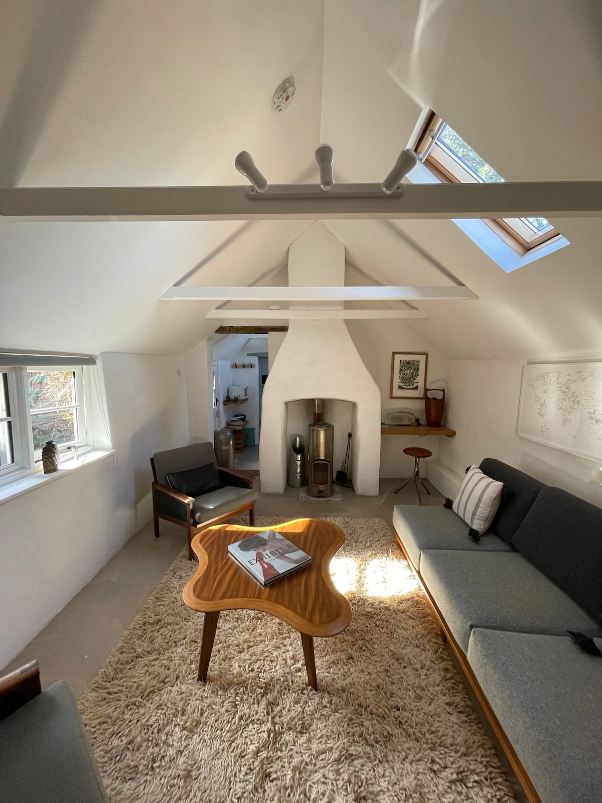 A living area features light-colored walls and a sloped ceiling with skylights allowing natural light to fill the space. A cozy grey sofa and two armchairs are arranged around a wooden coffee table, which sits atop a soft area rug. A sleek fireplace is positioned against one wall.
