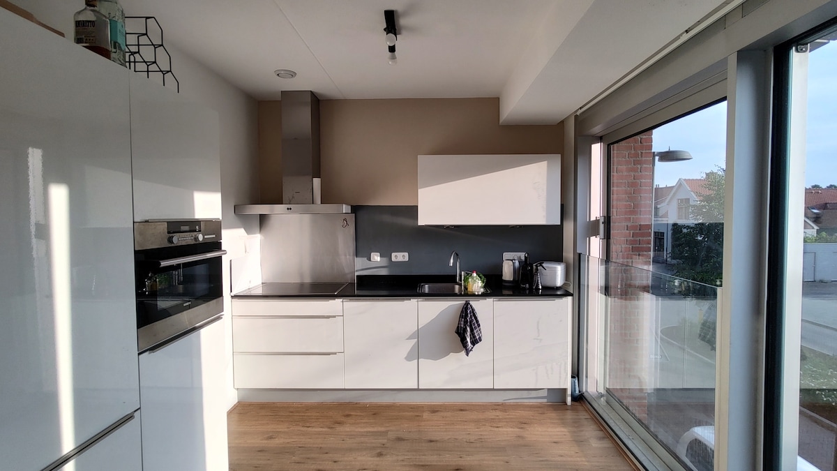 A modern kitchen space features sleek white cabinets and countertops, with stainless steel appliances. A large window allows natural light to fill the room, while a view of the outside area is visible. A towel hangs neatly on one of the cabinet handles.