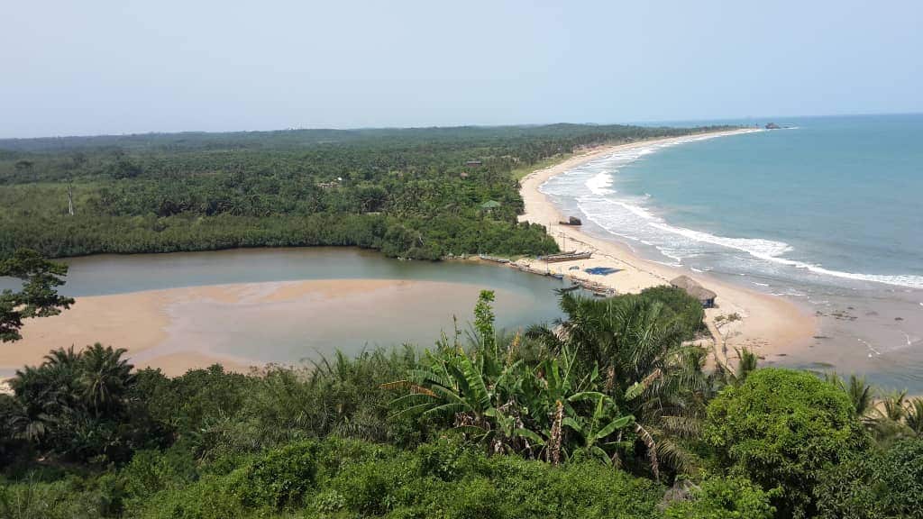 A panoramic view of the coastline is presented, featuring a wide sandy beach curving along the shoreline. Lush greenery borders the beach area, and a calm lagoon lies in the foreground, reflecting the surrounding landscape and clear blue sky.
