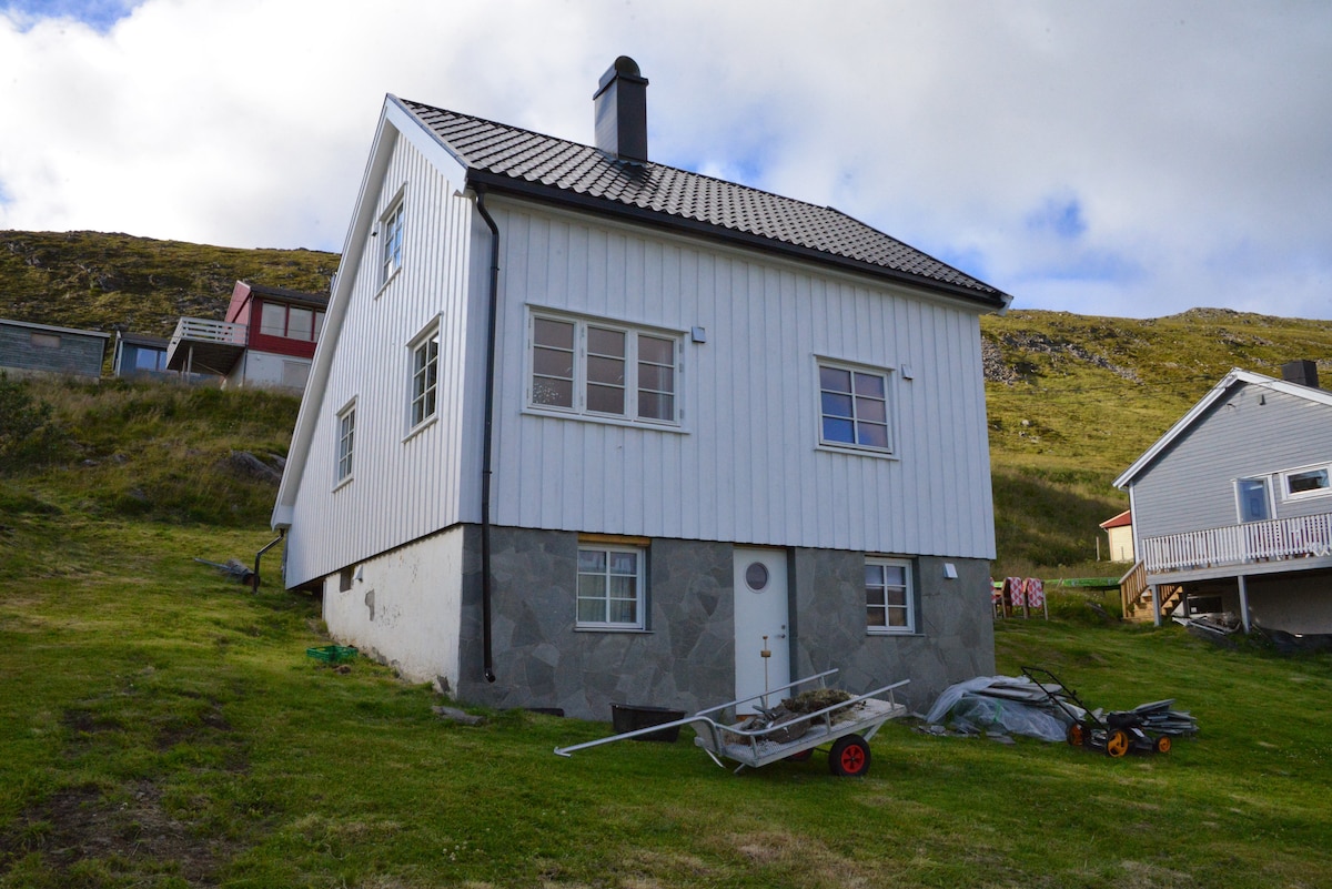 A two-story white house is situated on a grassy slope, showcasing a modern design with a sloped roof. The stone foundation contrasts with the wooden upper section. Nearby, various outdoor items, including wheelbarrows and garden tools, are visible, surrounded by the natural landscape.