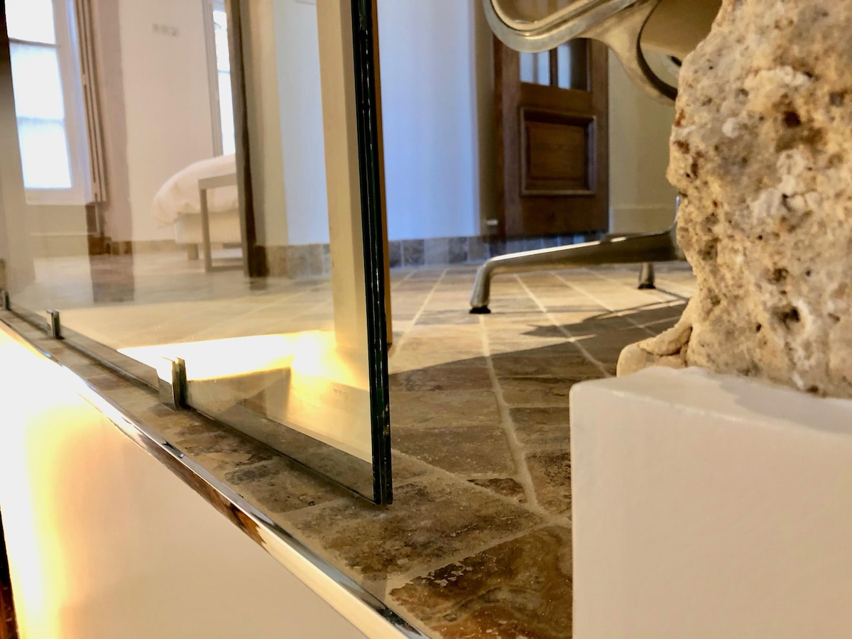 A glass partition separates the modern shower area from a spacious bathroom floor, which features natural stone tiles. A textured wall can be seen, contributing to the overall design. A glimpse of the bedroom is visible in the background, showcasing a minimalistic style.