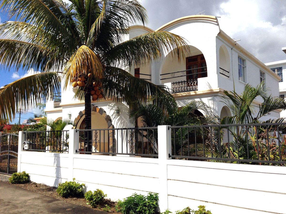 The villa presents a charming exterior with white walls and classic architectural arches. Lush palm trees frame the entrance, while vibrant tropical plants decorate the surrounding landscape. A gated fence adds privacy and security to the welcoming facade.