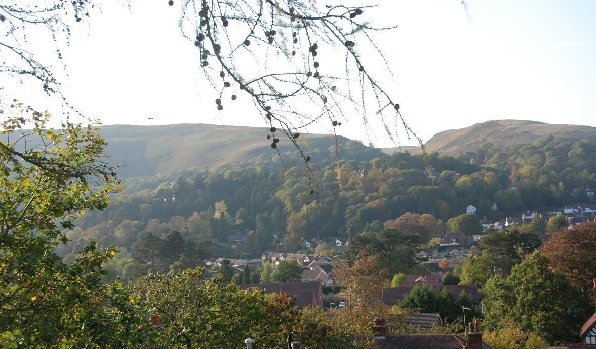 A scenic view captures the town of Church Stretton nestled among rolling hills. Lush greenery covers the slopes in the background, while residential buildings are visible below, creating a harmonious blend with the natural landscape.
