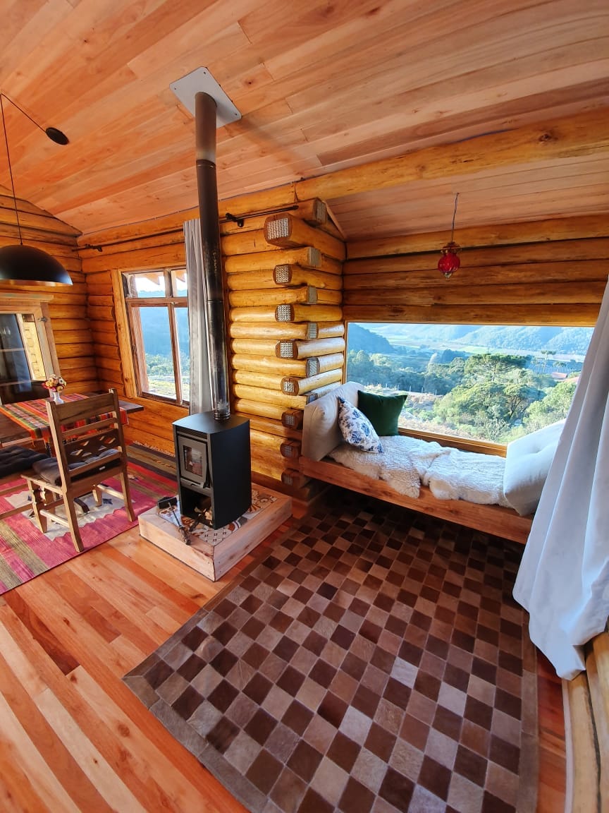 A cozy seating area is situated by a large window, offering sweeping views of the surrounding landscape. A small dining table with chairs is present, alongside a wood-burning stove. Rich wooden logs create a warm atmosphere, complemented by a patterned rug on the wooden floor.