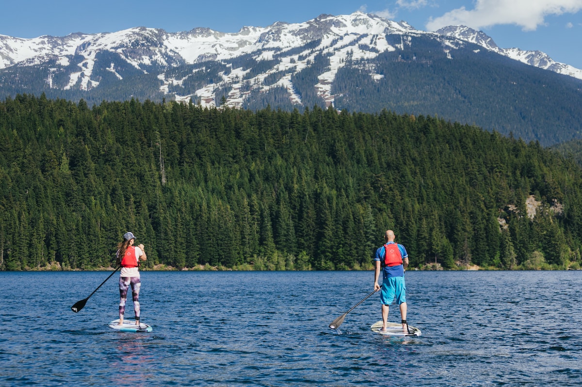 Two individuals are seen paddle boarding on a calm lake, with a backdrop of snow-capped mountains and dense evergreen forests. Their vibrant life jackets contrast against the blue water, emphasizing the serenity of the natural setting.