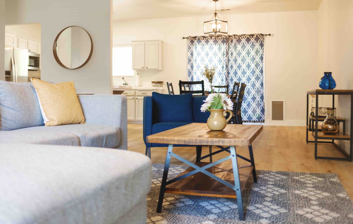 A cozy living area features a light gray couch and a blue accent chair, arranged around a wooden coffee table. Natural light fills the space, highlighting the patterned area rug beneath. The dining area is visible in the background, complemented by white cabinetry and a geometric-patterned curtain.