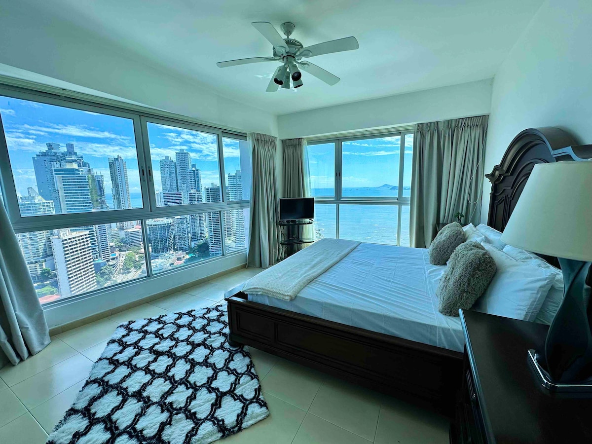 A spacious bedroom features a king-sized bed with neutral bedding, complemented by decorative pillows. Large windows provide panoramic views of Panama City and the bay, and a flat-screen TV is visible on a stand. A plush area rug adds texture to the room.