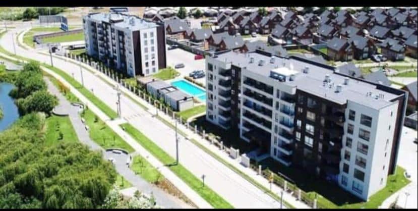 Aerial view of a modern condominium complex, showcasing multiple buildings surrounded by landscaped greenery. A swimming pool is visible in the courtyard area. A nearby road is seen connecting to residential homes and pathways, creating a sense of community and accessibility.