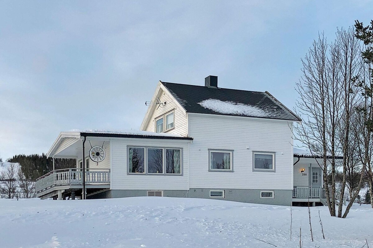 The two-story holiday home features a white exterior and a dark roof, surrounded by a snowy landscape. Large windows allow natural light to fill the interior. A covered terrace extends from the house, providing an outdoor space for relaxation. Sparse trees dot the surroundings.