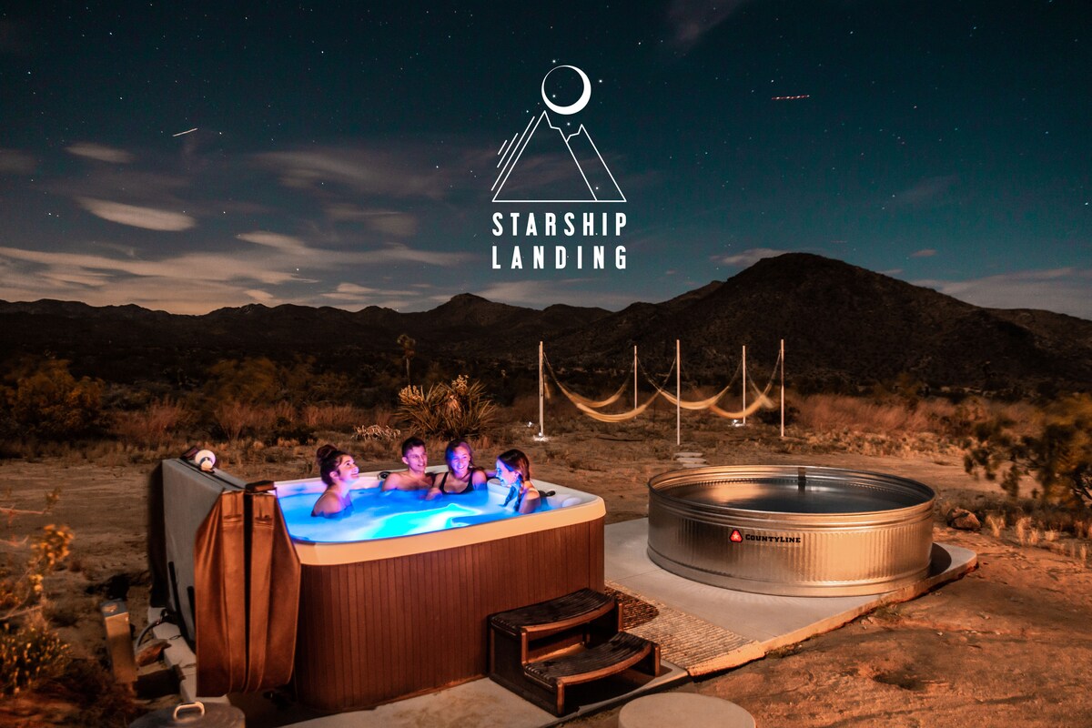 A hot tub glows invitingly under the starry sky, surrounded by a serene desert landscape. Adjacent to the hot tub, a cowboy tub pool reflects the surrounding mountains. Safety hammocks are set nearby, enhancing the outdoor relaxation experience.