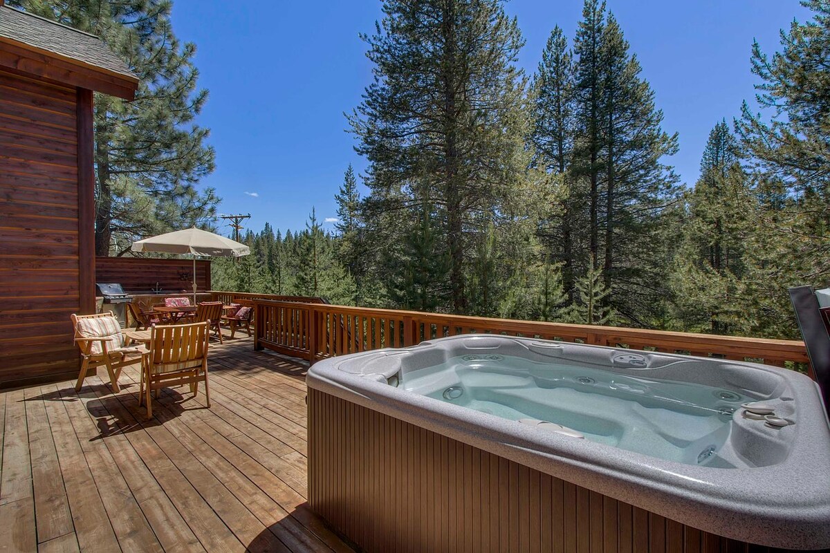 A spacious deck is shown with a hot tub positioned near the edge, surrounded by tall trees. Wooden lounge chairs and a table provide outdoor seating, while a large umbrella offers shade for relaxation.