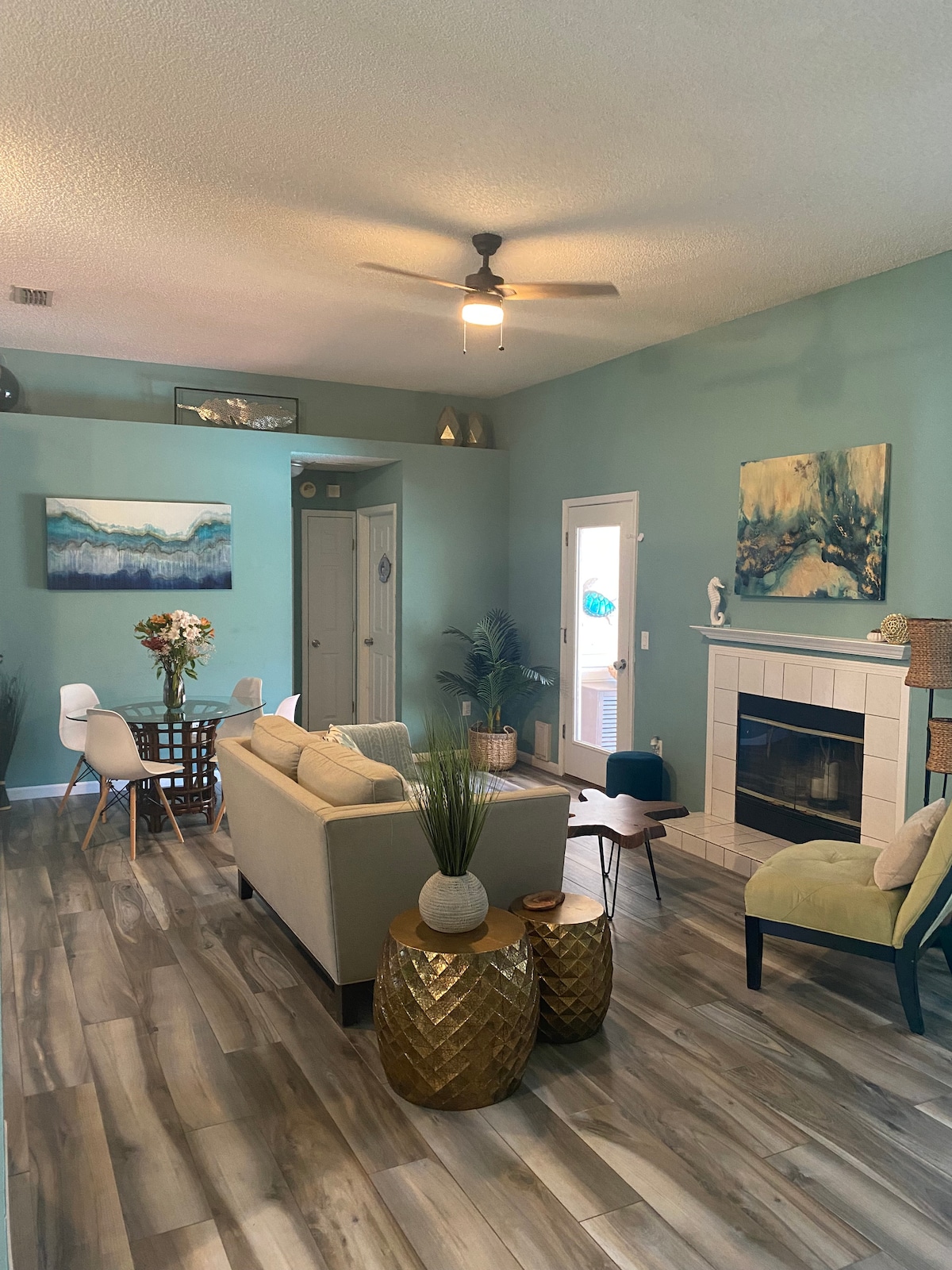 A welcoming living space features light blue walls and ample natural light. A spacious couch is complemented by a round dining table for four. Decorative elements include a fireplace and plants, creating an inviting atmosphere for relaxation.