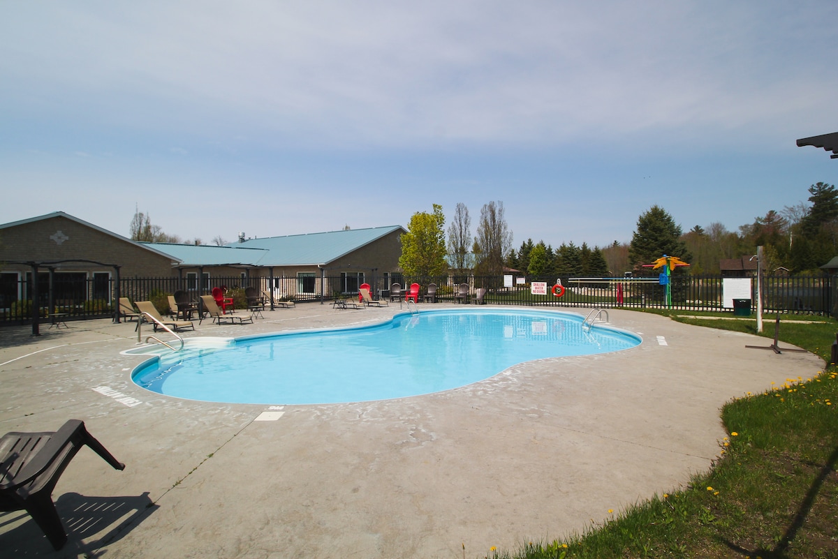 An outdoor swimming pool is featured, surrounded by a concrete deck with lounge chairs positioned for sunbathing. Lush greenery is visible in the background, along with buildings providing shade. A separate play area with colorful equipment can be seen nearby.