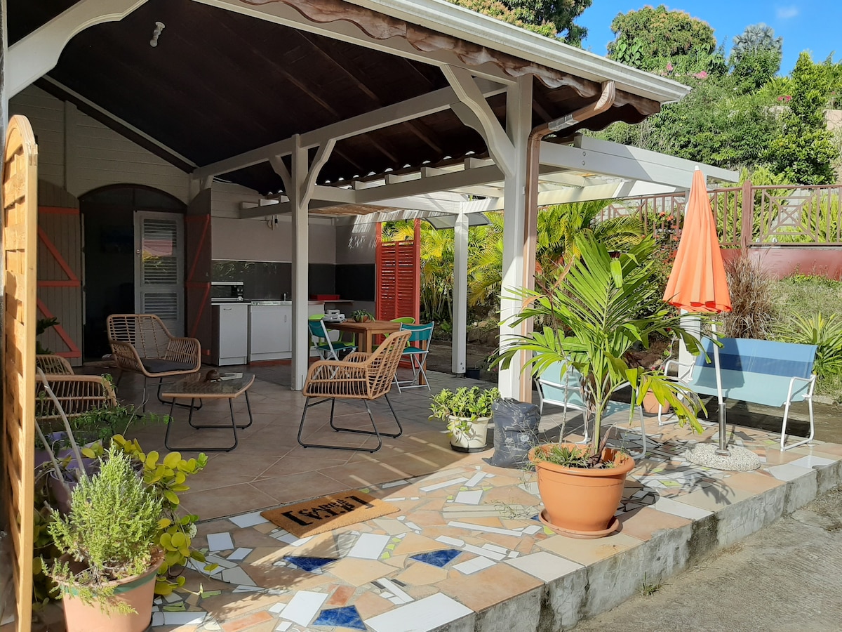 An outdoor terrace features a comfortable seating area with woven chairs and bright orange umbrellas. A kitchenette can be seen in the background, surrounded by lush greenery and colorful planters. The patterned tiled flooring adds a touch of character to the space.