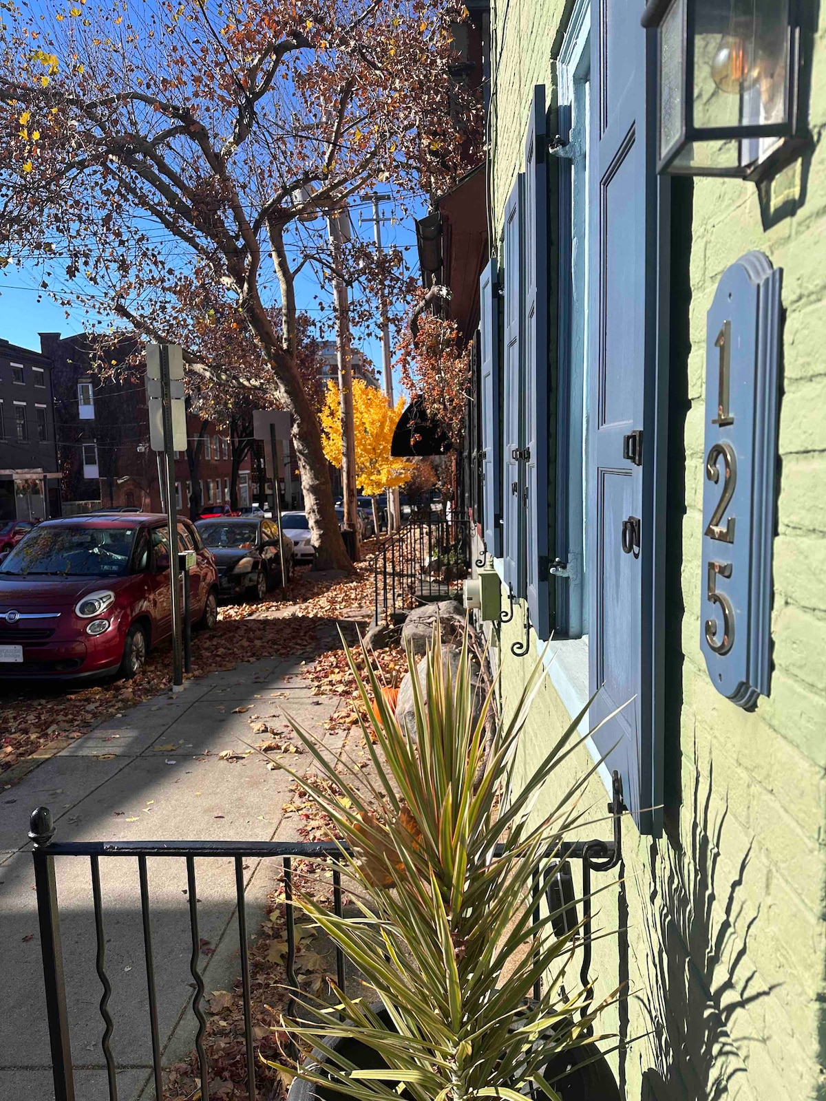 The front of the house features vibrant green exterior walls and blue shutters. A decorative fence lines the walkway adorned with fallen leaves. A number '125' is displayed prominently beside the doorway. Nearby, a young tree showcases golden autumn leaves.