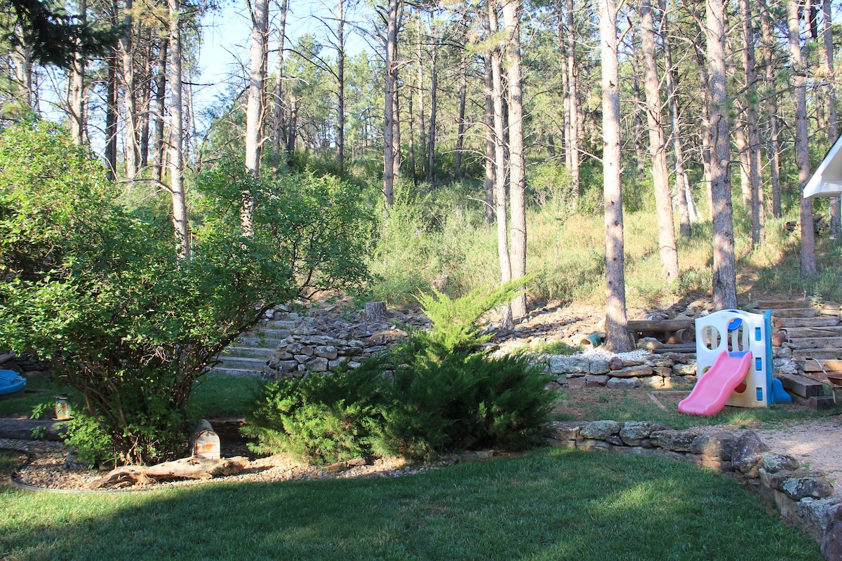A serene backyard is framed by tall trees, providing a peaceful atmosphere. A small play structure with a pink slide is positioned nearby, surrounded by lush green grass and a variety of shrubs. Natural stone features and steps lead towards a wooded area.