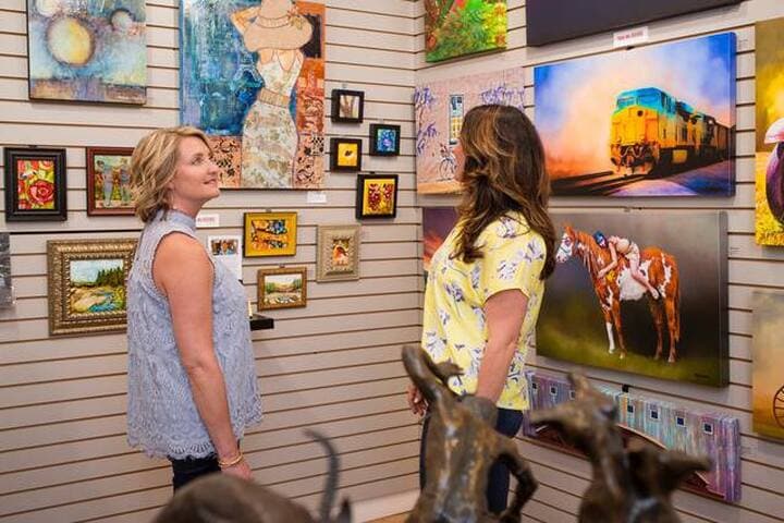 Two individuals are seen admiring various artworks displayed on a gallery wall. The collection features colorful paintings, including landscapes and animal portraits, framed in different styles. Sculptures of dogs are positioned in the foreground, adding to the artistic ambiance of the setting.