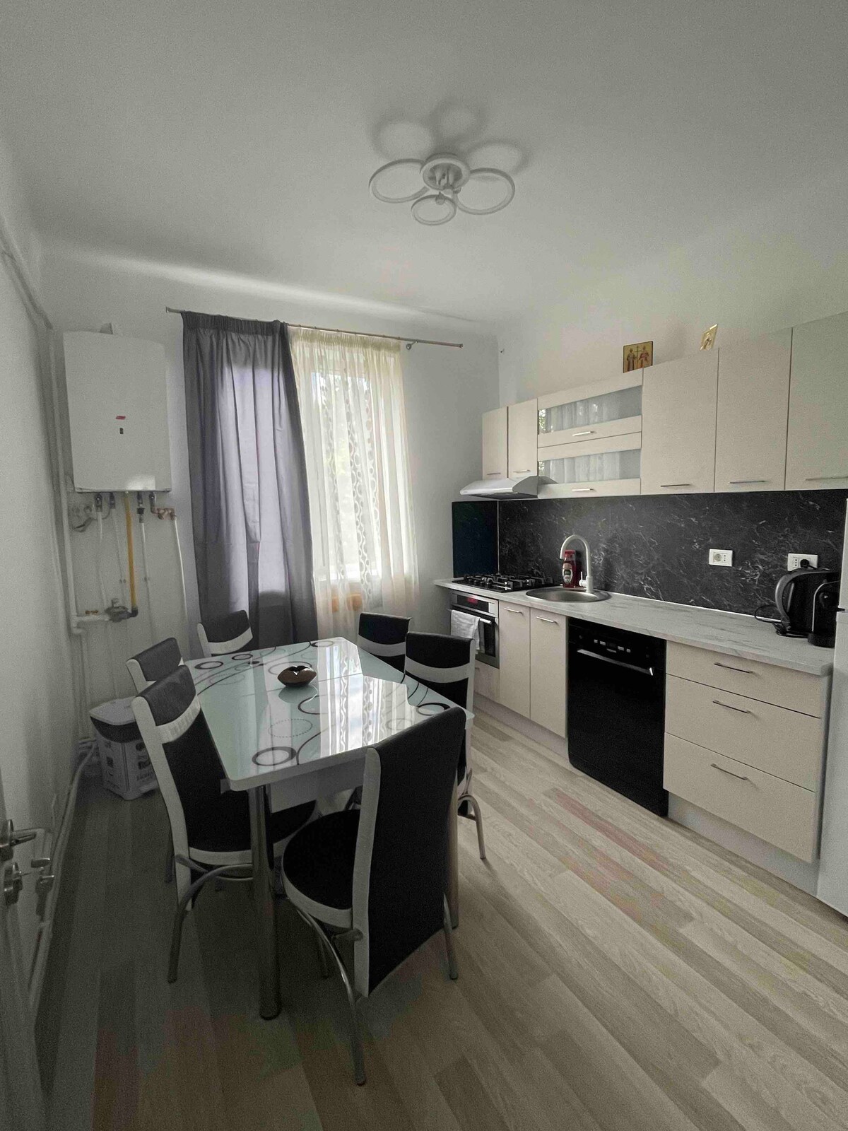 A modern kitchen featuring a sleek black countertop and an array of stainless steel appliances. A rectangular glass dining table is surrounded by six chairs. Natural light enters through sheer curtains, brightening the space, while cabinetry provides ample storage.
