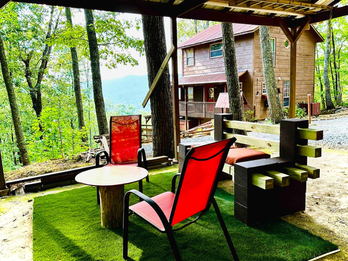 An outdoor seating area is featured under a covered patio, surrounded by trees and offering a view of the distant mountains. Two red chairs and a wooden bench are arranged around a circular table set on a grassy surface.