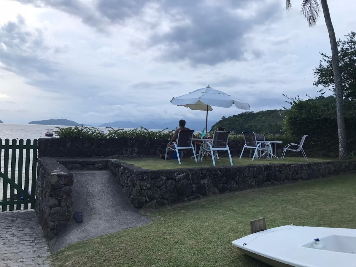A grassy area features a stone wall and outdoor seating, including a table and chairs beneath a large umbrella. The view extends towards the water and nearby hills, creating an open and inviting space suited for relaxation.