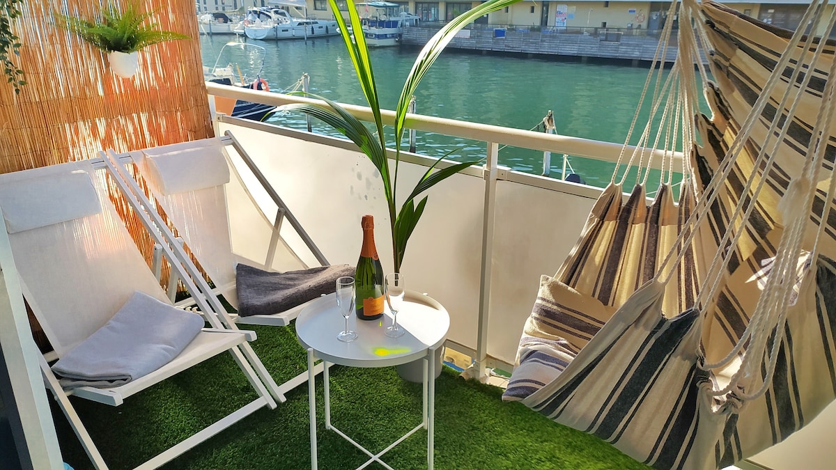 The terrace features two reclining chairs and a striped hammock, creating a comfortable seating area next to a small table set with glasses and a bottle of champagne. Lush greenery and a view of the canal add tranquility to the space.