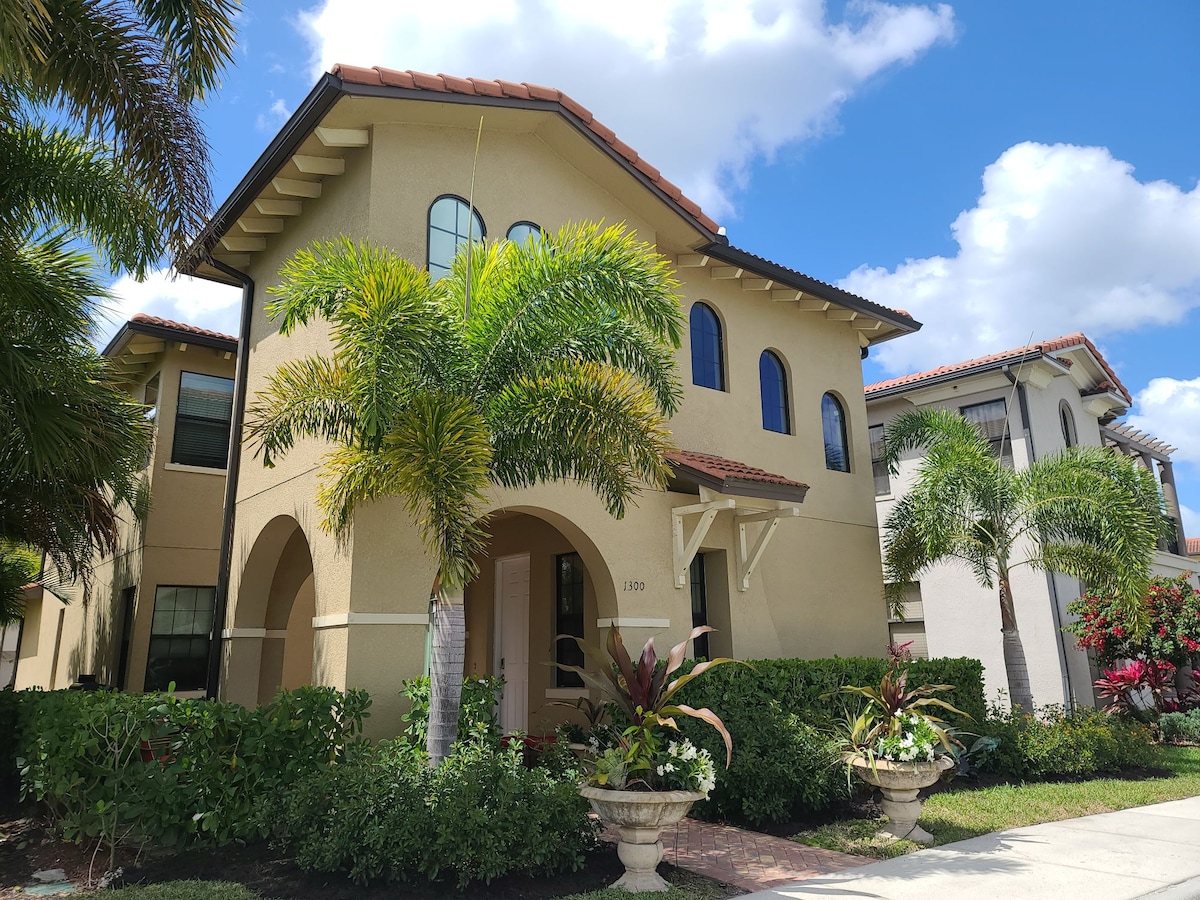 A two-story home with a Mediterranean style is framed by palm trees and lush landscaping. The exterior features arched windows and a warm-toned stucco facade, complemented by colorful flower pots at the entrance. The skies above are bright blue with scattered clouds.