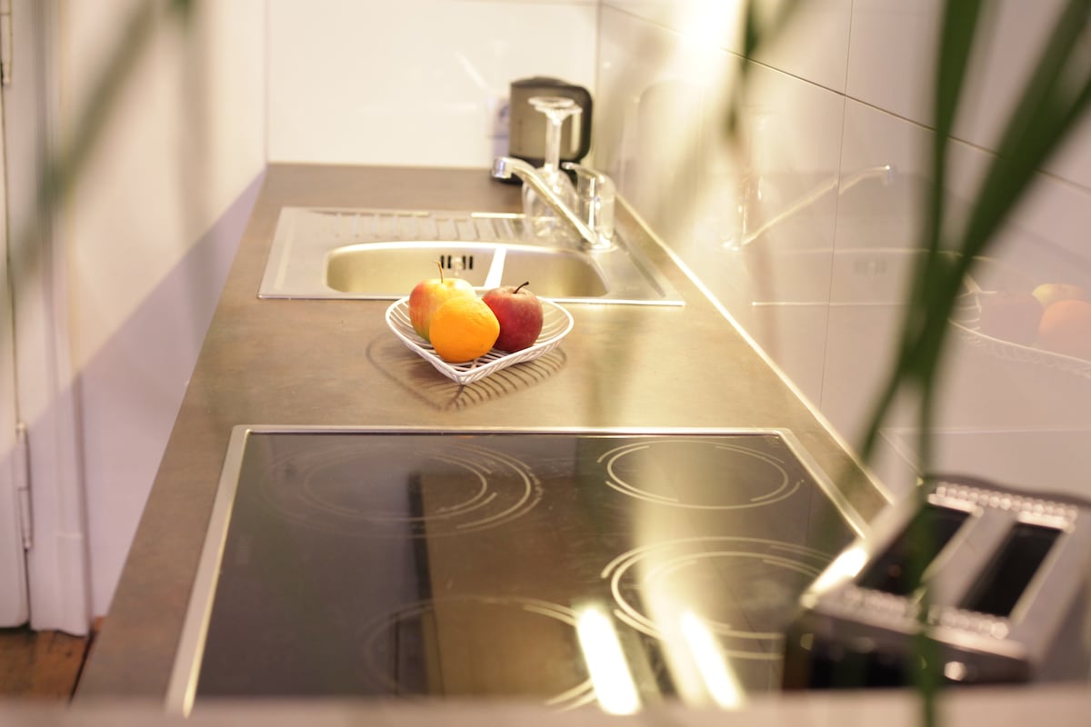 A modern kitchen counter is displayed, featuring a sleek stove and a sink. A bowl containing assorted fruits sits at the left side, while a toaster is positioned nearby. Reflections on the countertop create a polished appearance against the backdrop of minimalist white tiles.
