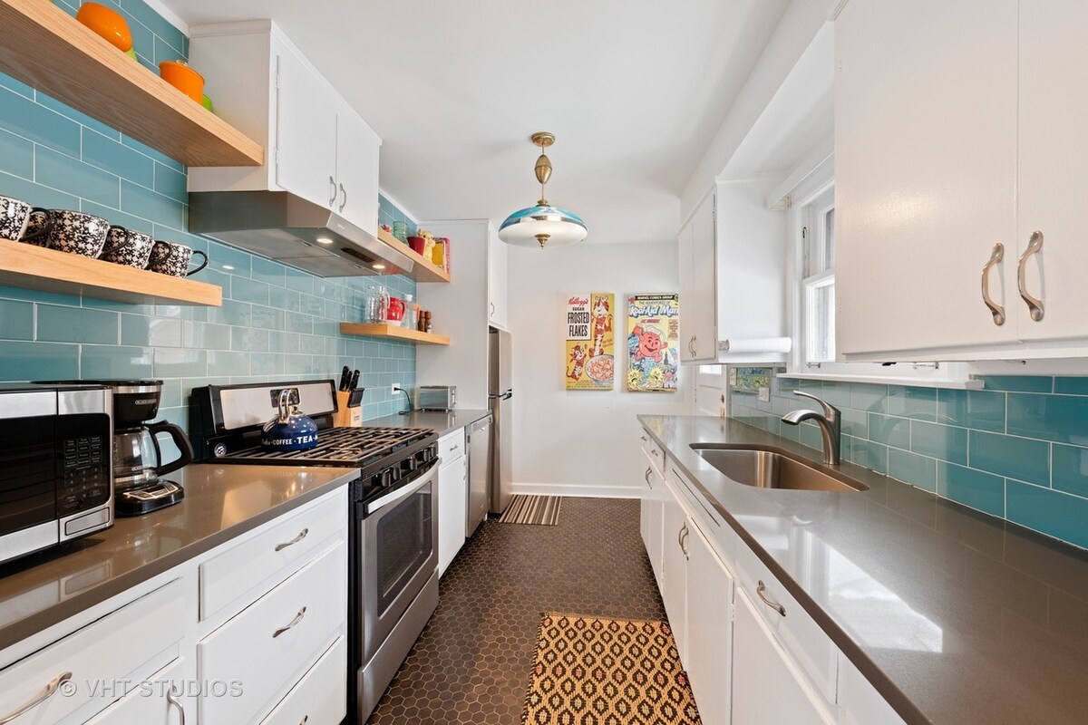 A modern kitchen features white cabinetry and a gray countertop. The walls are accented with teal tiles, and colorful artwork is displayed. Appliances include a microwave and a stove, with shelving that holds various dishware. A small rug is positioned in front of the sink.