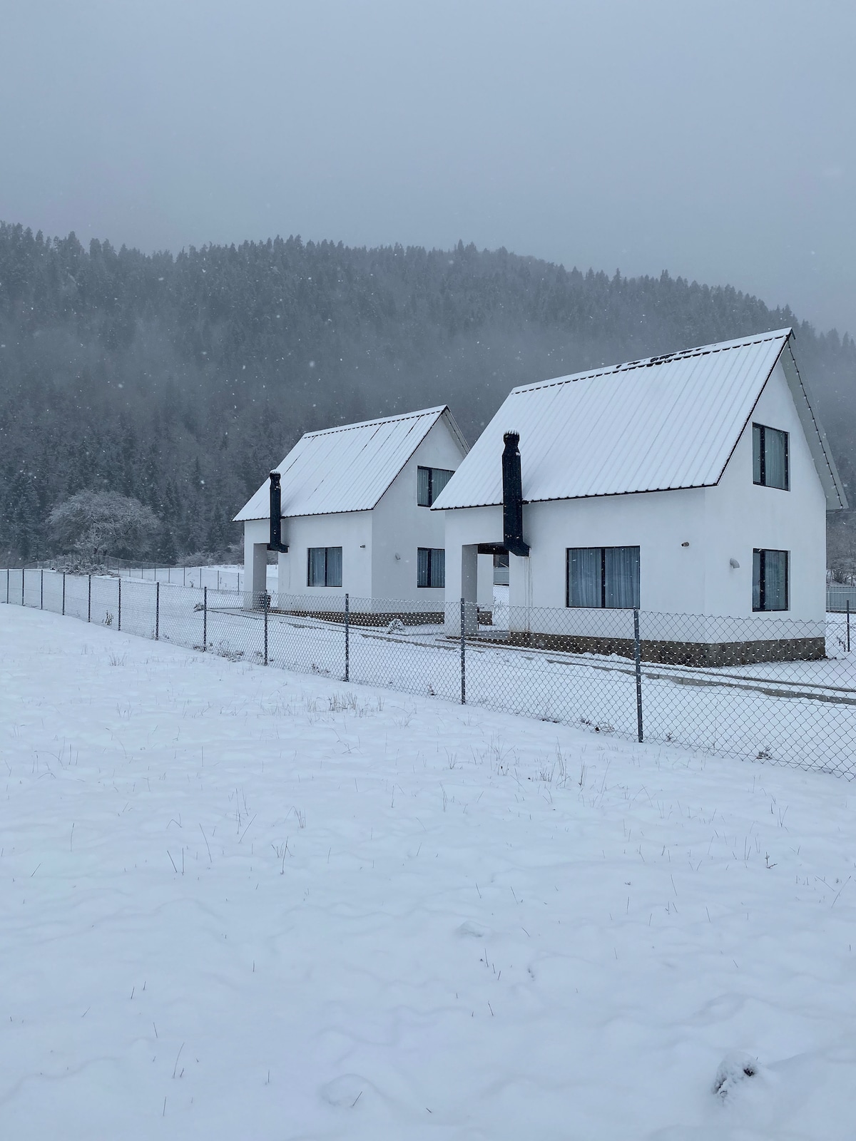 Two white houses with sloped roofs are situated in a snowy landscape. The surrounding area is peaceful, with forested hills in the background. A fence encloses the properties, creating a sense of privacy. Snow falls gently, adding to the serene winter atmosphere.