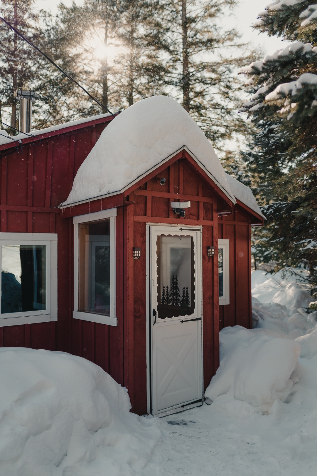 The entrance of the cabin is framed by snow-covered wooden walls and a peaked roof. A decorative door with a forest motif is centered, flanked by two windows. Sunlight filters through the trees, creating a serene winter ambiance.