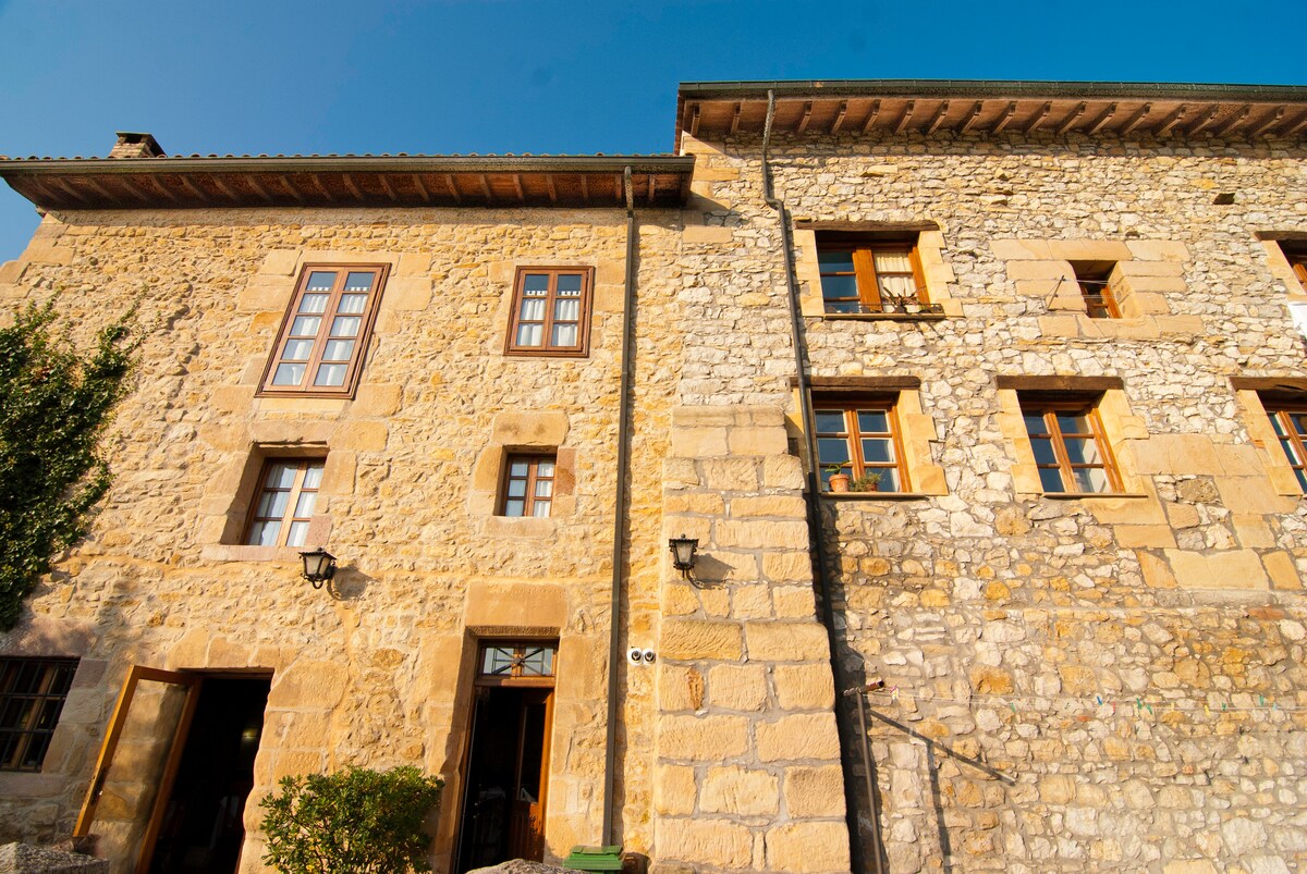 The exterior of a restored 18th-century building is visible, featuring a combination of natural stone and wooden elements. Multiple windows are aligned on both sides, showcasing the original architectural details. A blue sky provides a bright backdrop, enhancing the structure's historic charm.