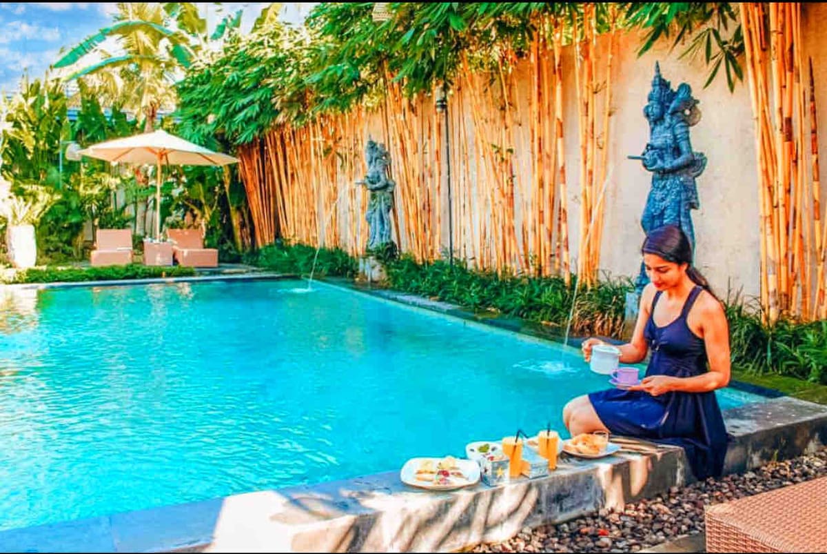 A tranquil pool area is surrounded by lush greenery and tall bamboo. A seated figure enjoys breakfast by the water, with a plate of food and drinks on a nearby surface. Two decorative statues enhance the serene ambiance of this outdoor space.