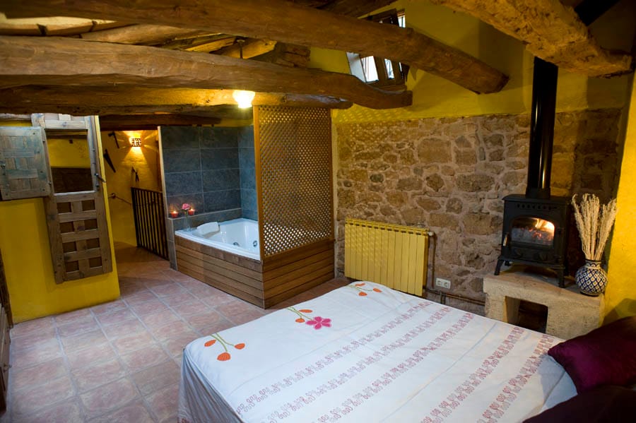 A warm, intimate space features a cozy bed adorned with a floral pattern. A private jacuzzi is situated nearby, complemented by wooden accents and stone walls. A small fireplace adds to the ambiance, while exposed beams highlight the rustic charm of the attic-style room.