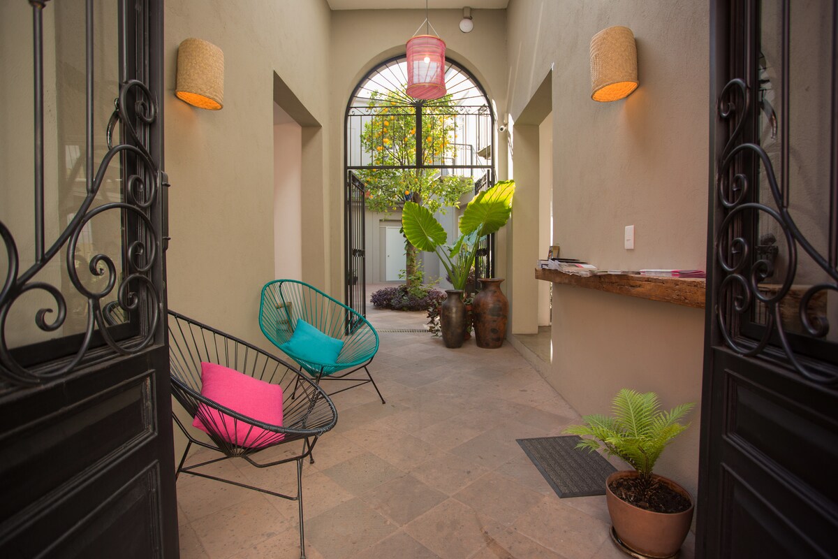 An inviting entrance area features two modern chairs in vibrant colors, complemented by potted plants and warm lighting. The open layout leads to a courtyard visible through the archway, enhancing the space's natural appeal.