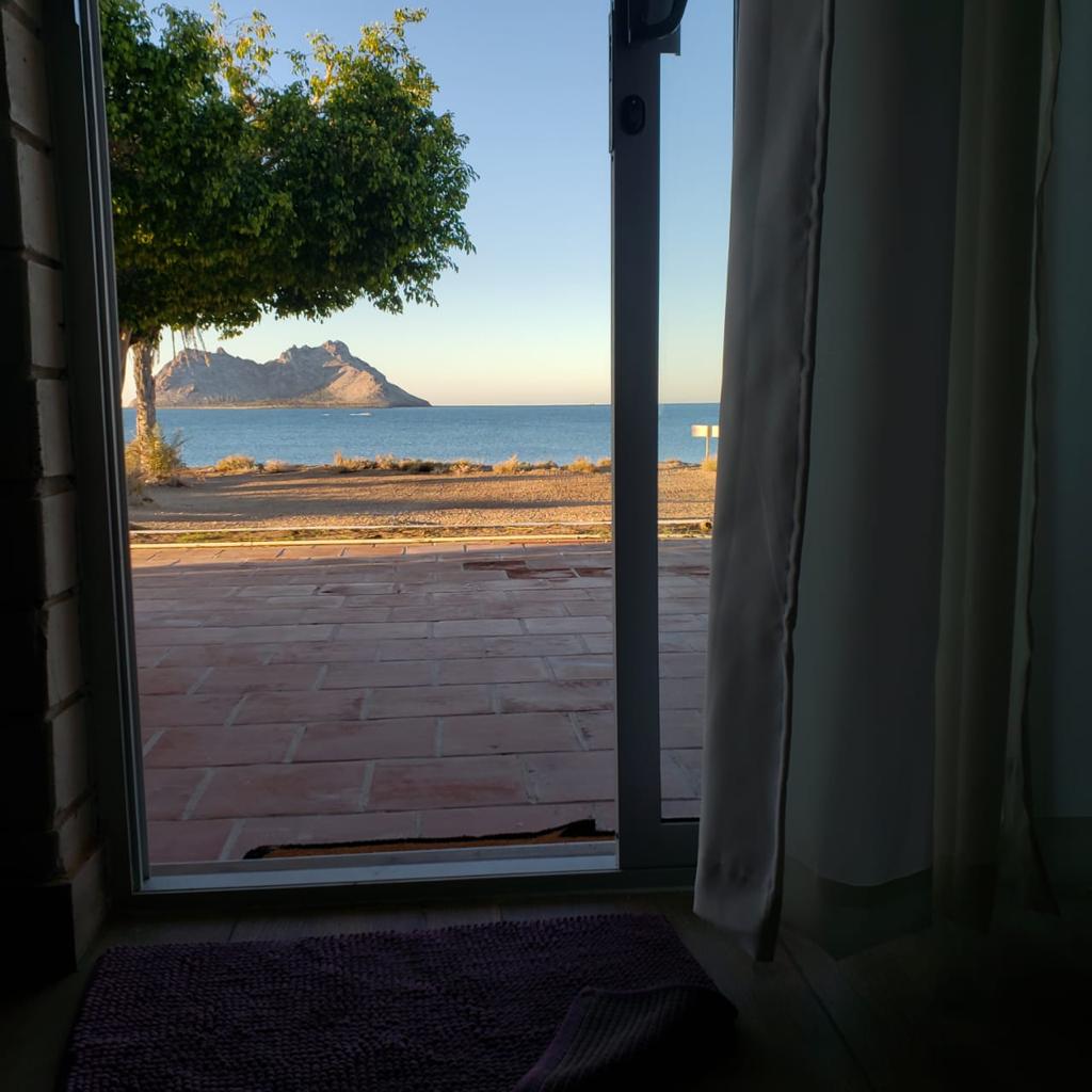 A view from the doorway captures the tranquil ocean and distant mountains under a clear sky. Light filters through sheer curtains, framing the outdoor scenery. The patio area is visible, featuring tiled flooring and well-maintained greenery along the coastline.